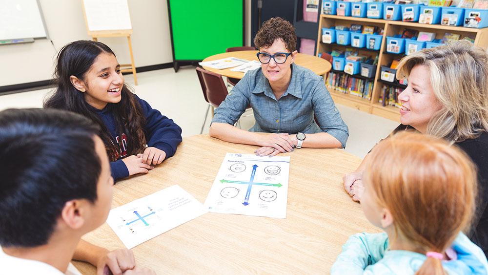 Sue Ducau sitting at table with elementary students