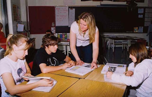 teacher leaning over desk speaking with students that are reading textbooks
