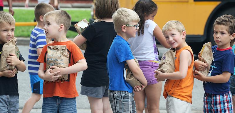 young elementary students standing together  near school bus holding paper bags