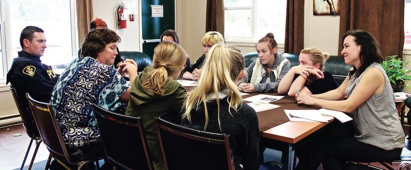 young teens, adults and a police officer sitting at boardroom table having discussion