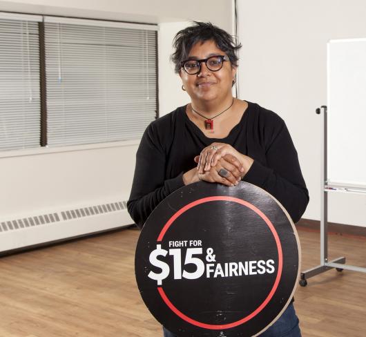 Deena Ladd standing in class holding $15 & Fairness logo