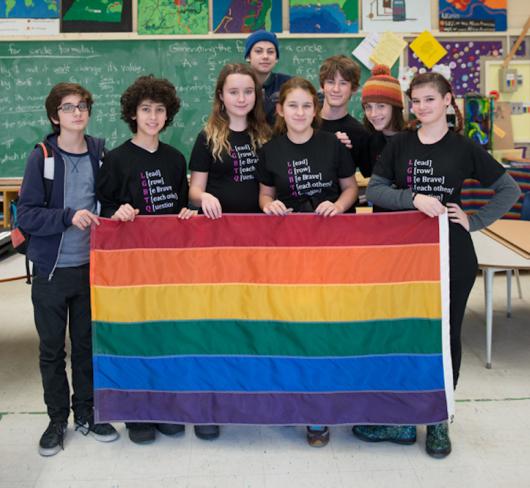 Students standing in classroom holding a rainbow flag