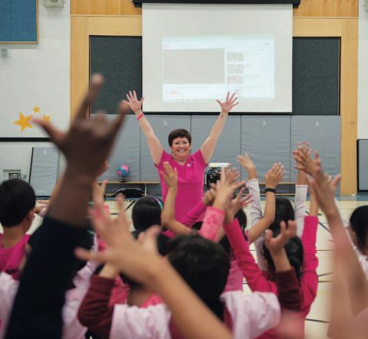 students in gymnasium mimicking teacher who has her hands in the air