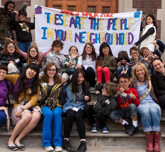 students sitting together holding sign that reads "These are people in our neighbourhood"