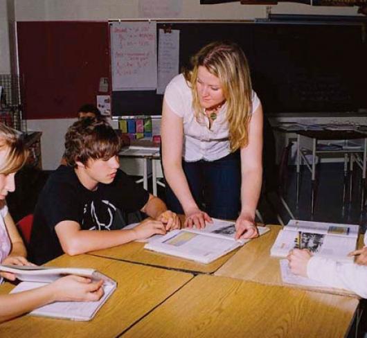 teacher leaning over desk speaking with students that are reading textbooks